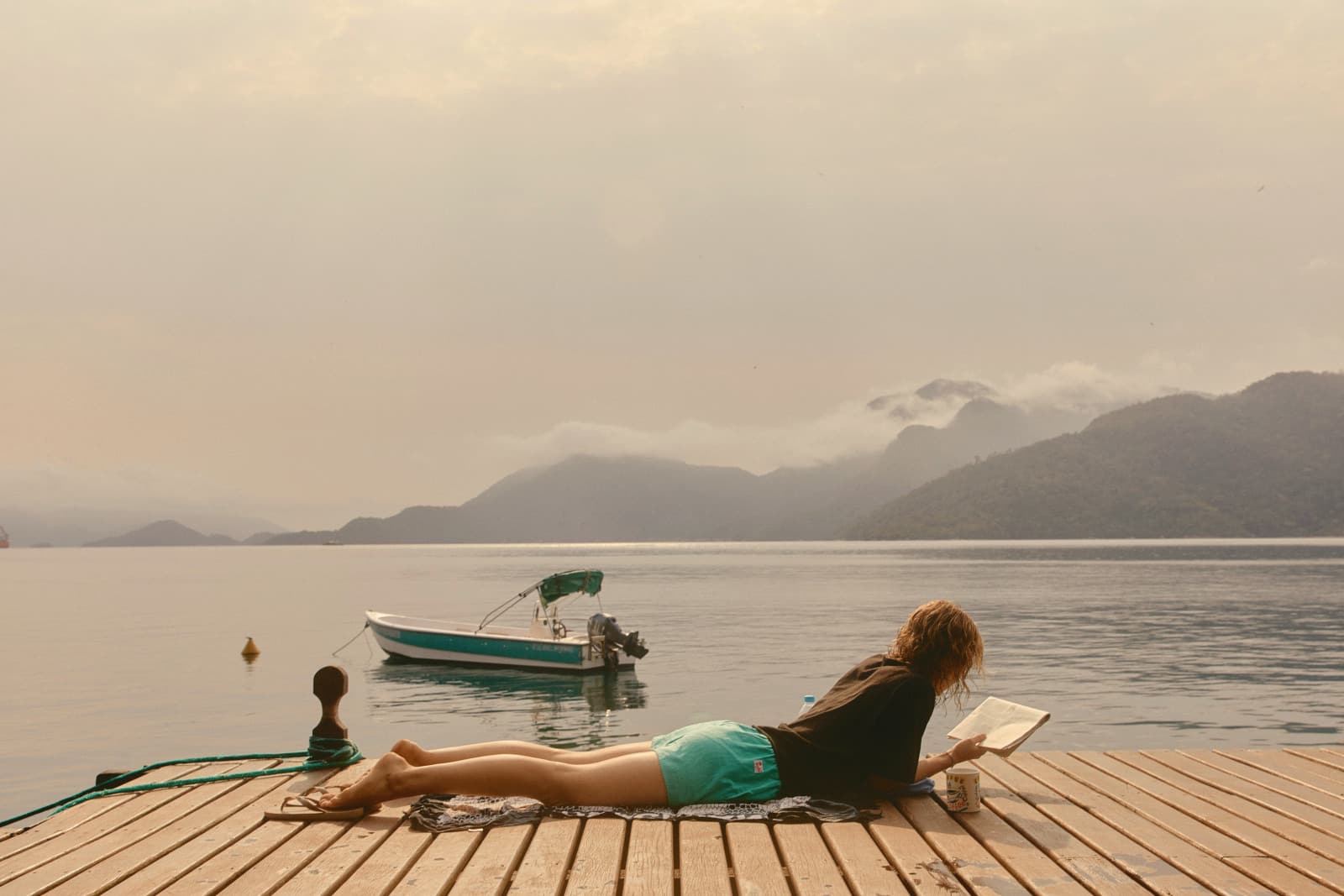 Someone lying on a jetty reading a book, a small boat moored in a calm bay below misty mountains